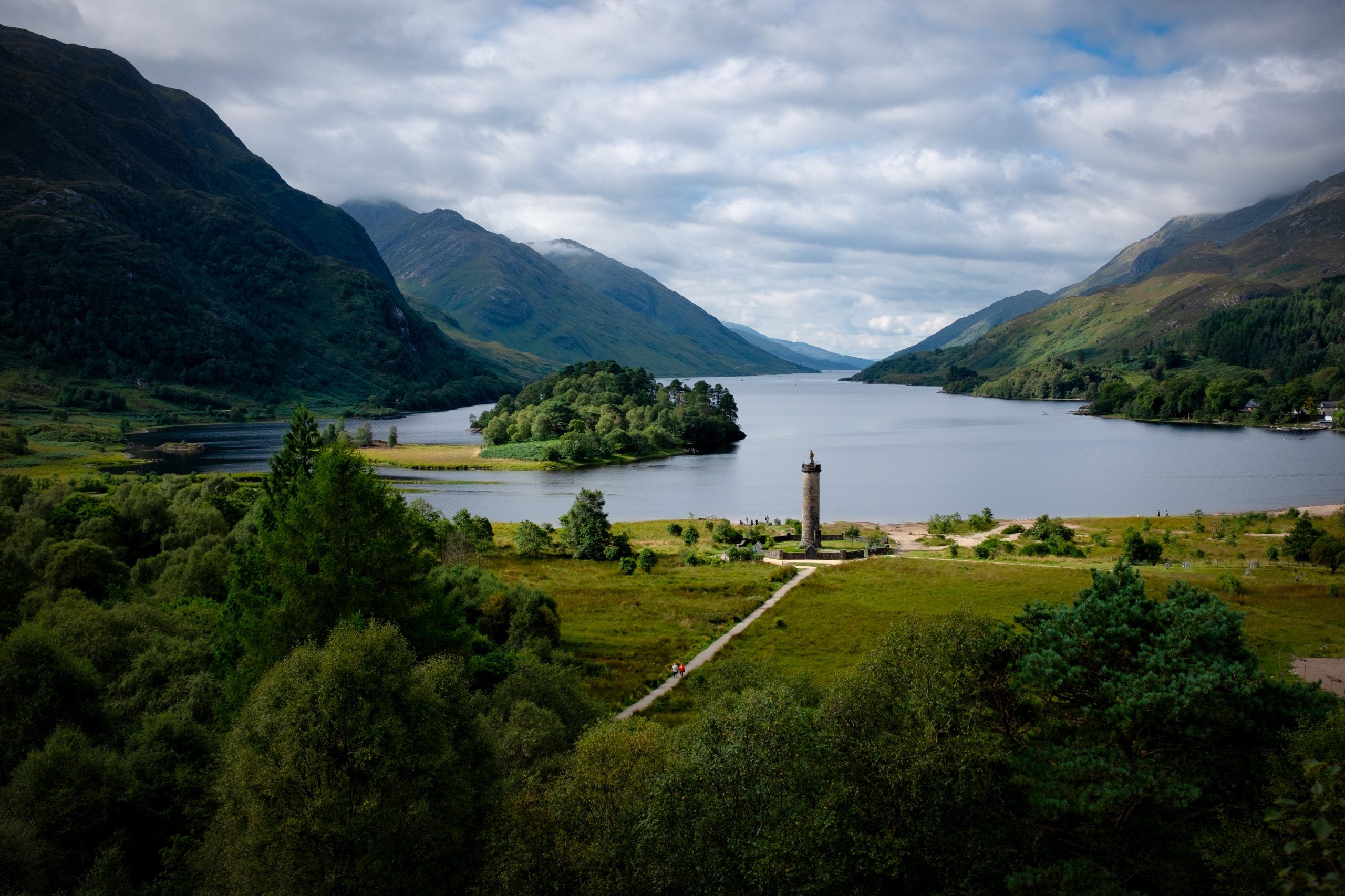A tall stone monument with a large loch or lake in the background. The lake is surrounded by mountains.